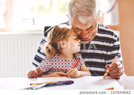 Cute little baby toddler girl and handsome senior grandfather painting with colorful pencils at home. Grandchild and man having fun together Cute little baby toddler girl and handsome senior grandfather painting with colorful pencils at home. Grandchild and man having fun together 107644682
