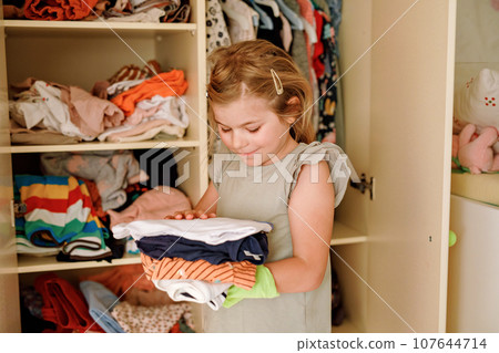 Little school girl standing by wardrobe with clothes. Preschool child putting shirts away and making decision for shirt to wear. Children get dressed in the morning for school. 107644714