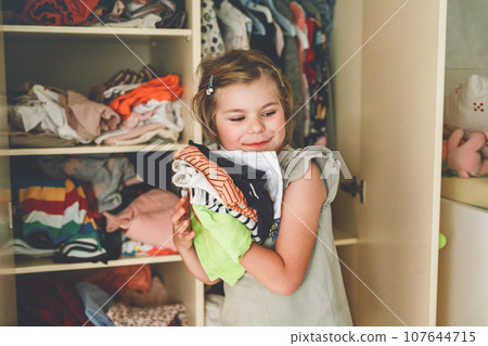 Little school girl standing by wardrobe with clothes. Preschool child putting shirts away and making decision for shirt to wear. Children get dressed in the morning for school. Little school girl standing by wardrobe with clothes. Preschool child putting shirts away and making decision for shirt to wear. Children get dressed in the morning for school. 107644715