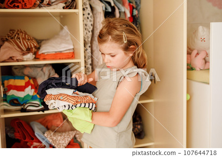 Little school girl standing by wardrobe with clothes. Preschool child putting shirts away and making decision for shirt to wear. Children get dressed in the morning for school. 107644718