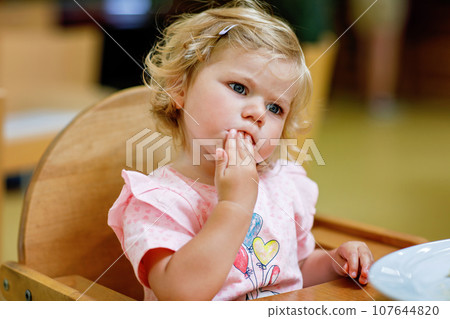 Adorable toddler girl eating healthy vegetables and unhealthy french fries potatoes. Cute happy baby child taking food from dish at daycare or nursery canteen. 107644820