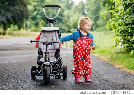 Cute adorable toddler girl sitting on pushing bicycle or tricycle. Little baby child going for a walk with parents cold cloudy day. Happy healthy kid in colorful clothes Cute adorable toddler girl sitting on pushing bicycle or tricycle. Little baby child going for a walk with parents cold cloudy day. Happy healthy kid in colorful clothes 107644871