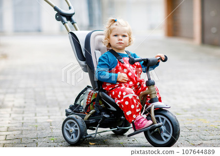 Cute adorable toddler girl sitting on pushing bicycle or tricycle. Little baby child going for a walk with parents cold cloudy day. Happy healthy kid in colorful clothes 107644889