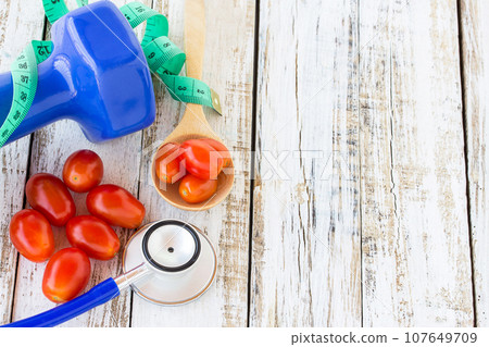 Blue stethoscope and dumbbell with red tomatos on white wooden table Blue stethoscope and dumbbell with red tomatos on white wooden table 107649709