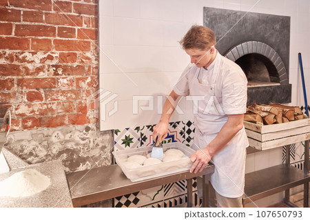 The chef prepare pizza. Raw pizza ready to bake. Cook in a apron in the kitchen Rolls out pizza dough. boxes for food delivery on background. 107650793