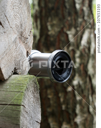 The camera lens peeks around the corner of a wooden, log wall, the photographer hides to take an unexpected photo. Secret work of a photographer, paparazzi. 107653193