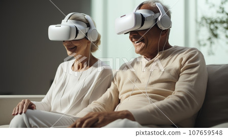 Senior mature couple wearing virtual reality headsets sits on the sofa having fun together. Happy retired people using modern VR goggle glasses. New trends and technology concept for all ages. 107659548