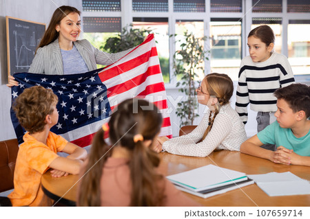 Group of curious preteen learners and young female teacher with flag of USA in classroom of academy 107659714