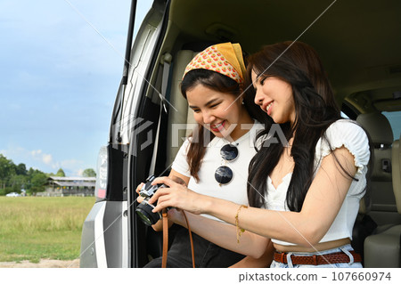 Cheerful women travelers sitting in opened trunk of car and checking pictures on camera. Road trip, traveling and lifestyle concept Cheerful women travelers sitting in opened trunk of car and checking pictures on camera. Road trip, traveling and lifestyle concept 107660974