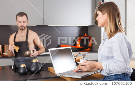 Woman with laptop while husband preparing food Woman with laptop while husband preparing food 107661100