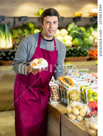 Male seller holding packaged carrots and sliced papaya standing in fruit and vegetable section of supermarket Male seller holding packaged carrots and sliced papaya standing in fruit and vegetable section of supermarket 107661494