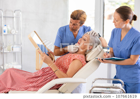 Man dermatologist examines skin of senior womans face, female nurse records data in client card 107661619