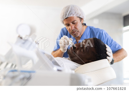 Male cosmetologist performs skin cleansing procedure for African man client with beauty machine Male cosmetologist performs skin cleansing procedure for African man client with beauty machine 107661828