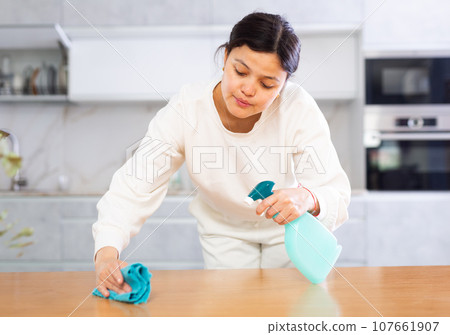 Smiling young woman wiping kitchen countertop with cleaning spray 107661907