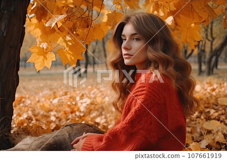 a woman in an orange sweater sitting on the ground with autumn leaves around her she is looking into the camera 107661919