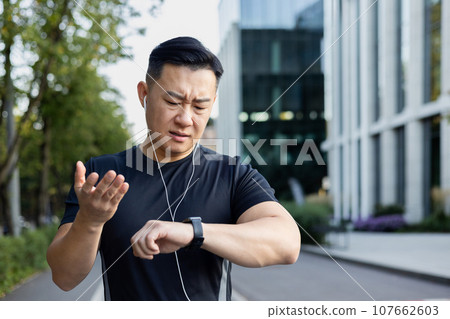Close-up photo. A young Asian man does sports, runs on a city street in headphones, looks sadly at the result on a smart watch, throws his hands in frustration. 107662603