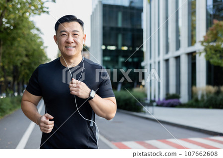 Portrait of an Asian young man running in headphones on a city street in the morning, smiling at the camera. 107662608