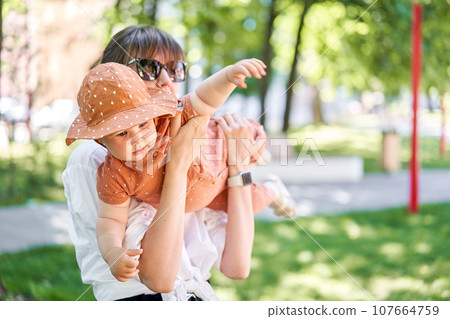 Portraits young mother and her daughter of acute 10 months old baby girl. Walking on the playground, on a sunny summer day. 107664759
