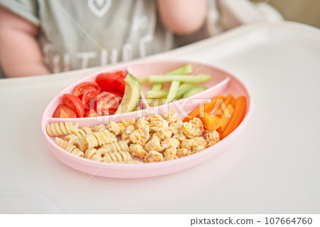Cute child eats healthy food pasta and vegetables steamed,. Portraits of a cute 10 months old baby girl. The baby sitting in a special high chair for babies. Cute child eats healthy food pasta and vegetables steamed,. Portraits of a cute 10 months old baby girl. The baby sitting in a special high chair for babies. 107664760