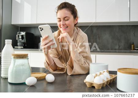 Attractive young cheerful girl baking at the kitchen, making dough, holding recipe book, having ideas Attractive young cheerful girl baking at the kitchen, making dough, holding recipe book, having ideas 107665237