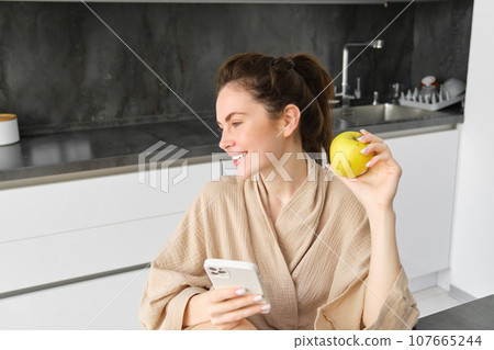 Image of attractive young woman in bathrobe, sits in kitchen, eats green apple and looks at mobile phone, uses smartphone app, orders food on application 107665244