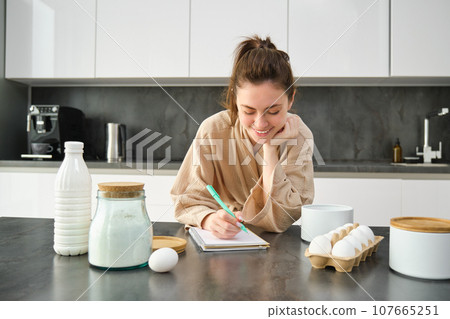 Attractive young cheerful girl baking at the kitchen, making dough, holding recipe book, having ideas Attractive young cheerful girl baking at the kitchen, making dough, holding recipe book, having ideas 107665251