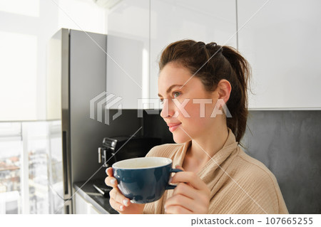 Close up portrait of beautiful young woman, posing in the kitchen, looking dreamy and thoughtful, contemplating morning with mug of coffee, waiting for breakfast 107665255