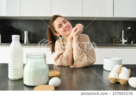 Attractive young cheerful girl baking at the kitchen, making dough, holding recipe book, having ideas 107665256