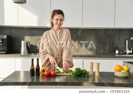 Portrait of good-looking woman cooking salad in the kitchen, chopping vegetables and smiling, preparing healthy meal, leading healthy lifestyle and eating raw food Portrait of good-looking woman cooking salad in the kitchen, chopping vegetables and smiling, preparing healthy meal, leading healthy lifestyle and eating raw food 107665265