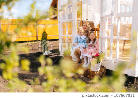 Remote view of elegance blonde young mother in dress sitting on doorstep of summer gazebo house with two little adorable daughters on sunny day, smiling looking at camera with happy expression. 107667031