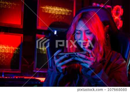 Gamer playing online game application on mobile phone wear gaming headphones, Asian woman live stream she play video game via smartphone at home neon lights living room Gamer playing online game application on mobile phone wear gaming headphones, Asian woman live stream she play video game via smartphone at home neon lights living room 107668080