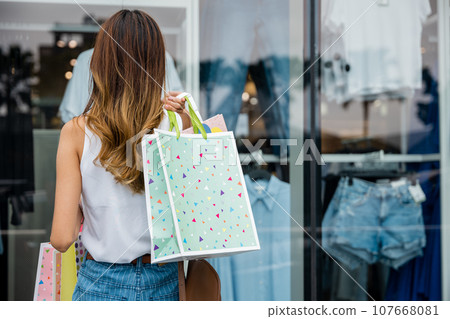 A happy customer carrying shopping bags stands in front of a store window, admiring the fashion display. She's excited about the sale and ready to spend on her favorite styles. A happy customer carrying shopping bags stands in front of a store window, admiring the fashion display. She's excited about the sale and ready to spend on her favorite styles. 107668081