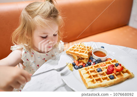 Happy 3 year old girl eat. Breakfast in cafe. Classic Viennese waffles with ice cream, berries and Maple syrup. Table in the restaurant. 107668092