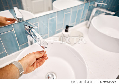 A man's hand touches the water in a beautiful sink with a metal faucet next to an mirror. Close-up of an elegant faucet in the bathroom sink next to stylish decorations.. High quality photo 107668096