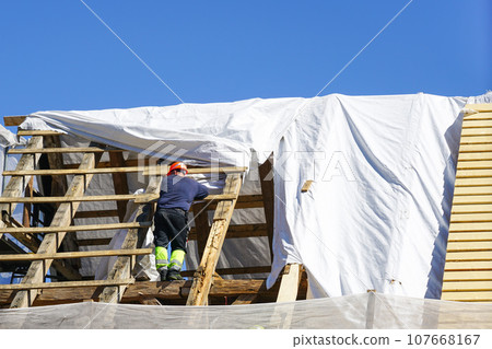 A worker replaces the damaged roof structures of an old wooden house, white safety net, blue sky A worker replaces the damaged roof structures of an old wooden house, white safety net, blue sky 107668167