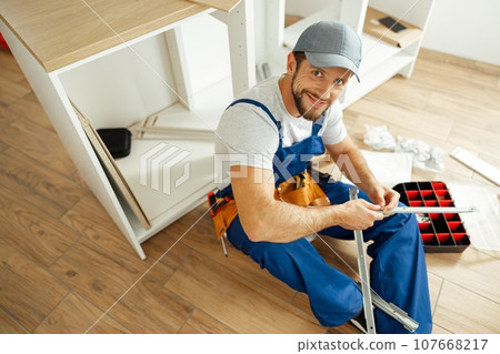 High angle view of handyman in uniform smiling at camera while sitting on the floor and assembling drawer in kitchen 107668217