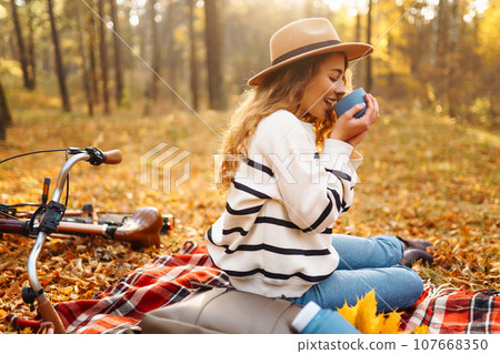 Hiker sitting on picnic blanket, drinking hot drink in autumn forest. Woman resting in nature. 107668350