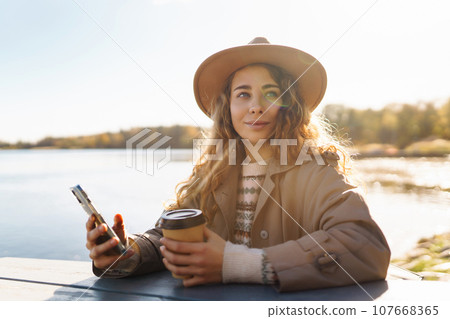 Smiling woman in hat on terrace with phone enjoys sunny autumn weather. Concept of relaxation, rest. 107668365
