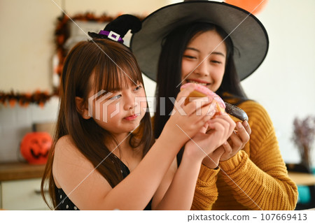 Happy mother and cute girl daughter are having fun preparing Halloween desserts in kitchen Happy mother and cute girl daughter are having fun preparing Halloween desserts in kitchen 107669413