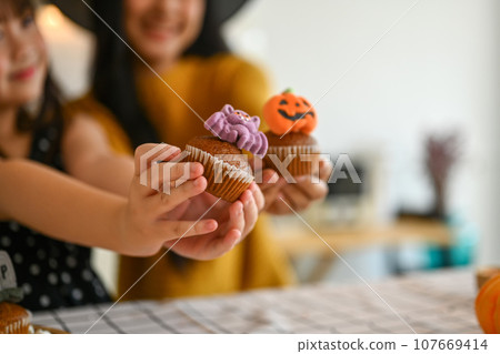 Close cup little girl and mother hands holding delicious Halloween cupcakes. Halloween celebration concept Close cup little girl and mother hands holding delicious Halloween cupcakes. Halloween celebration concept 107669414