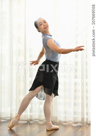 Portrait of elegant female dancer in pointe shoes practicing ballet positions near large window 107669508