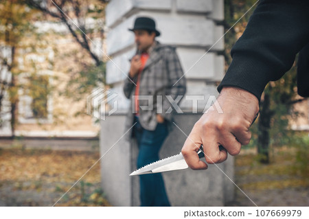 Hand of a robber with a knife on the background of a man in the park. Crime, violence concept 107669979
