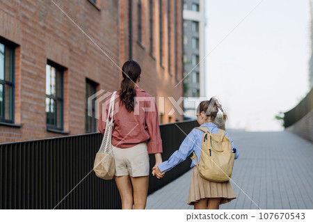 Mother and school girl with a backpack holding hands while going to school. Concept of back to school. Mother and school girl with a backpack holding hands while going to school. Concept of back to school. 107670543