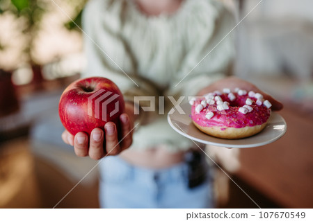 Diabetic woman is choosing between fresh apple and a sweet doughnut. Importance of proper nutrition and diet in diabetes. Diabetic woman is choosing between fresh apple and a sweet doughnut. Importance of proper nutrition and diet in diabetes. 107670549