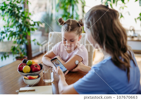 Girl with diabetes checking blood glucose level at home using continuous glucose monitor. Mother connects CGM to a smartphone to monitor her blood sugar levels in real time. 107670663