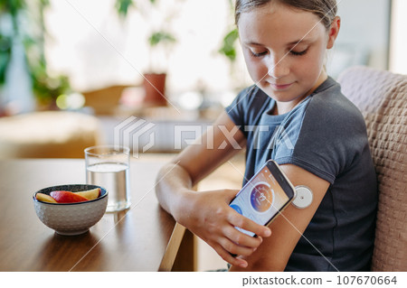 Girl with diabetes checking blood glucose level at home using continuous glucose monitor. Schoolgirl connecting CGM to smartphone, monitoring her blood sugar levels in real time. 107670664