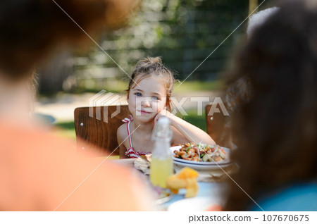 Beautiful little girl sitting at a table eating grilled food outdoors. The girl at family garden party. Beautiful little girl sitting at a table eating grilled food outdoors. The girl at family garden party. 107670675