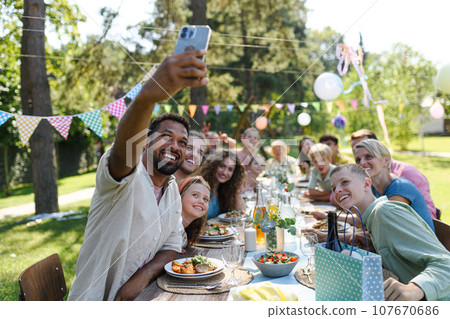 The whole family sitting at the table taking group selfie at the family garden party. A family gathering to celebrate birthday. 107670686