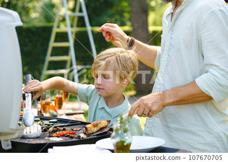 Young boy turning grilled cheese with tongs. Father and son grilling together at garden bbq party. Young boy turning grilled cheese with tongs. Father and son grilling together at garden bbq party. 107670705