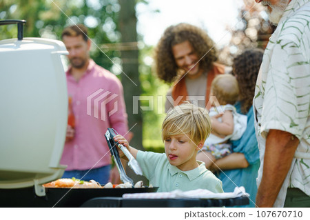 Boy turning grilled food with tongs. Grandfather and grandson are grilling together at a garden bbq party. Boy turning grilled food with tongs. Grandfather and grandson are grilling together at a garden bbq party. 107670710